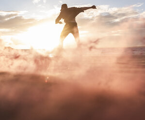Mann beim Bewegungstraining am Strand mit buntem Rauch bei Sonnenuntergang - UUF15059
