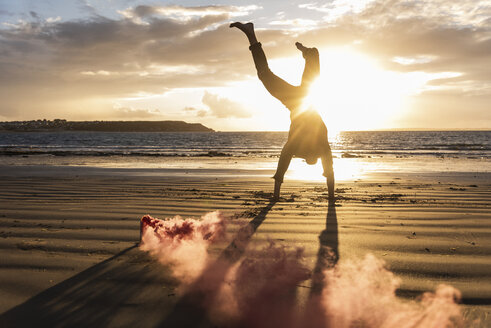 Man doing movement training at the beach with colorful smoke at sunset - UUF15064