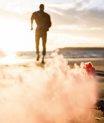 Junger Mann läuft am Strand bei Sonnenuntergang mit buntem Rauch - UUF15067