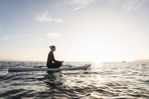 Junge Frau meditiert auf einem Paddelbrett bei Sonnenuntergang, lizenzfreies Stockfoto