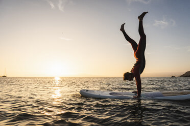 Junger Mann macht Handstand auf Paddleboard bei Sonnenuntergang - UUF15085