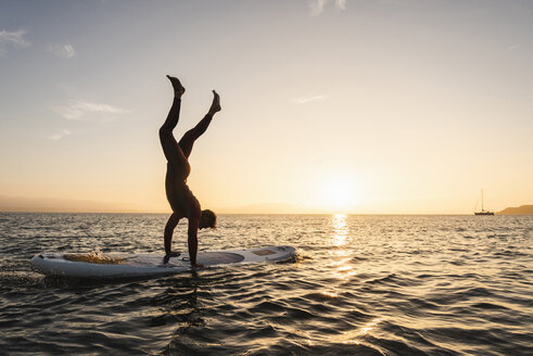 Junger Mann macht Handstand auf Paddleboard bei Sonnenuntergang - UUF15086