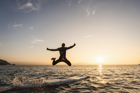 Junger Mann springt bei Sonnenuntergang vom Paddleboard ins Wasser - UUF15089