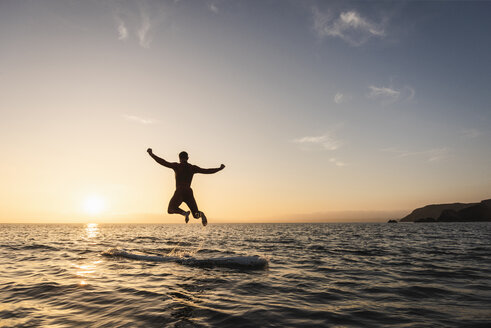 Junger Mann springt bei Sonnenuntergang vom Paddleboard ins Wasser - UUF15090