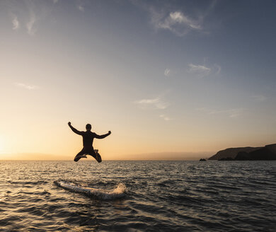 Junger Mann springt bei Sonnenuntergang vom Paddleboard ins Wasser - UUF15091