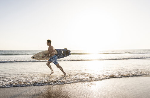 Junger Mann läuft am Strand, trägt Surfbrett - UUF15115