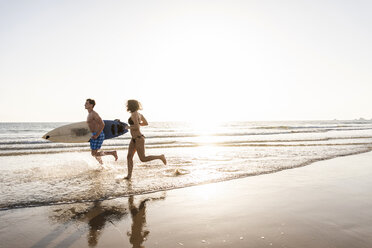 Young couple running on beach, carrying surfboard - UUF15122