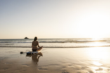 Junge Frau praktiziert Yoga am Strand, sitzt auf einem Surfbrett und meditiert - UUF15136