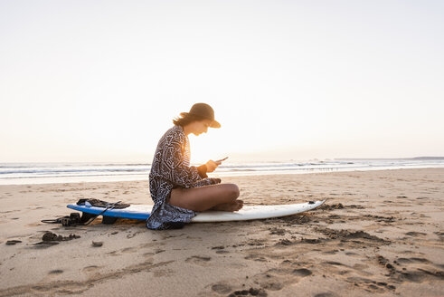 Junge Frau sitzt auf einem Surfbrett am Strand und benutzt ein Smartphone - UUF15159