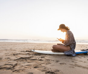 Young woman sitting on surfboard at the beach, using smartphone - UUF15160