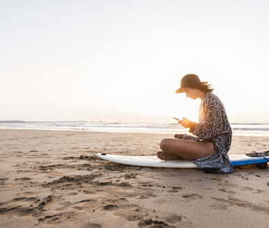 Junge Frau sitzt auf einem Surfbrett am Strand und benutzt ein Smartphone - UUF15160