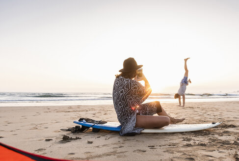 Junge Frau sitzt auf einem Surfbrett und fotografiert einen jungen Mann, der am Strand Handstand übt - UUF15161