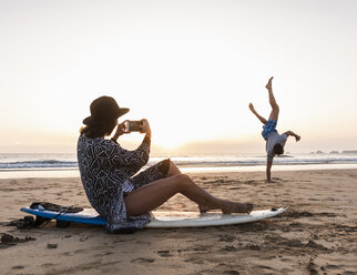 Junge Frau sitzt auf einem Surfbrett und fotografiert einen jungen Mann, der am Strand Handstand übt - UUF15163