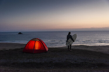 Young woman camping on the beach, carrying surfboard at night - UUF15166