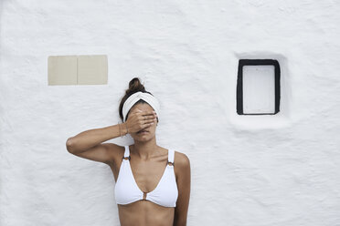 Junge Frau mit weißem Bikinioberteil lehnt an einer weißen Wand und bedeckt die Augen mit ihrer Hand - IGGF00592