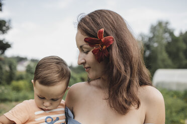 Mutter mit Blume im Haar hält Baby im Garten - KMKF00549