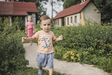 Baby boy walking in garden with sister in background - KMKF00557