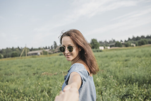 Smiling woman wearing sunglasses holding hand of partner in rural field - KMKF00564