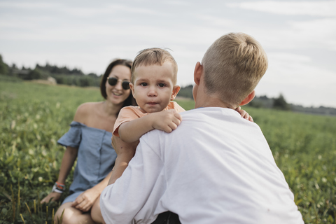 Bruder tröstet weinendes Baby auf einem Feld mit Mutter im Hintergrund, lizenzfreies Stockfoto