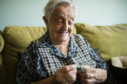 Porträt einer lächelnden älteren Frau, die zu Hause auf der Couch häkelt, lizenzfreies Stockfoto