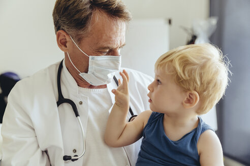 Toddler sitting on lap of pediatrician, wearing protective mask - MFF04476