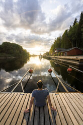 Finnland, Kajaani, Mann sitzt auf Steg und beobachtet Sonnenuntergang, Rückansicht - KKAF01709