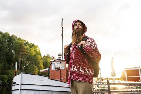 Finland, Kajaani, Cheerful young man. wearing houded jacket, standing on jetty, carrrying rucksack - KKAF01717
