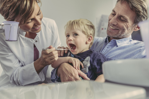 Eine Zahnärztin untersucht einen kleinen Jungen, der auf dem Schoß seines Vaters sitzt, lizenzfreies Stockfoto