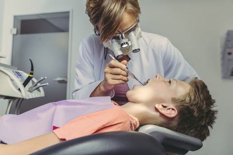 Zahnarzt, der die Zähne eines Jungen untersucht, mit Kopflupe und zahnärztlichem Instrument, lizenzfreies Stockfoto
