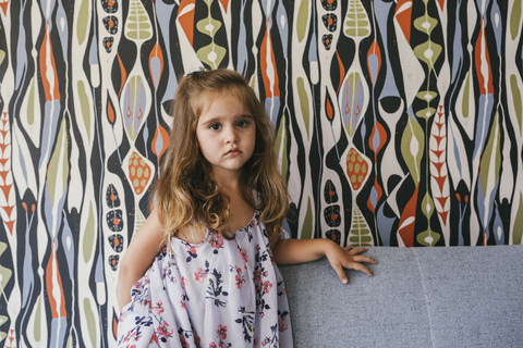 Porträt eines ernsten kleinen Mädchens auf einer Couch vor einer gemusterten Tapete, lizenzfreies Stockfoto