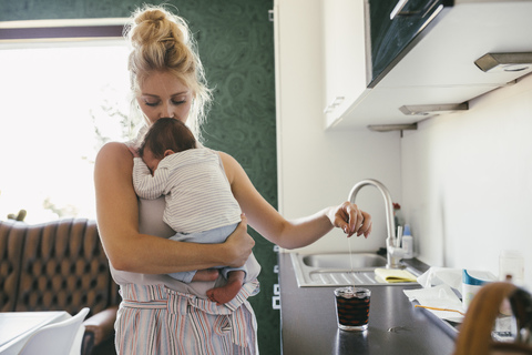 Mutter hält ihr neugeborenes Baby in der Küche und kocht Tee, lizenzfreies Stockfoto