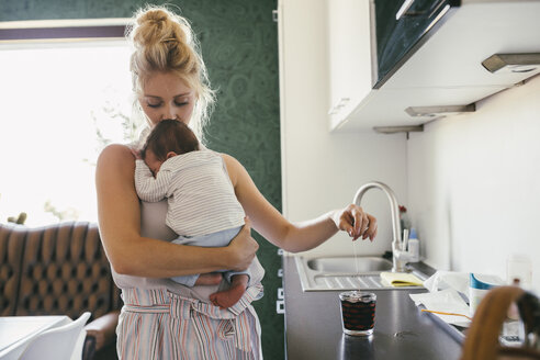 Mother holding newborn baby in kitchen while making tea - MFF04612