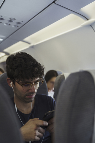 Junger Mann in einem Flugzeug mit Handy und Kopfhörern, lizenzfreies Stockfoto