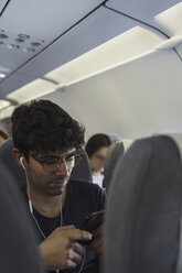 Junger Mann in einem Flugzeug mit Handy und Kopfhörern - KKAF01775