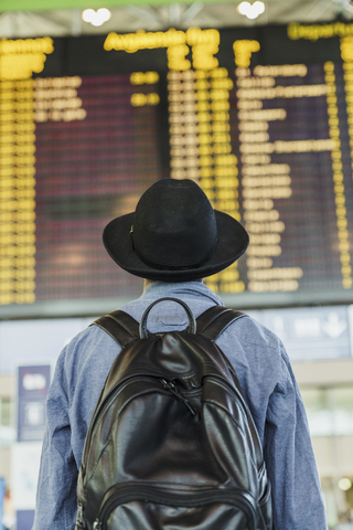 Junger Mann mit Hut und Rucksack schaut auf die Abflugtafel am Flughafen, lizenzfreies Stockfoto