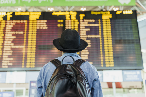 Junger Mann mit Hut und Rucksack schaut auf die Abflugtafel am Flughafen, lizenzfreies Stockfoto