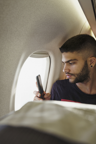 Junger Mann in einem Flugzeug mit Mobiltelefon, lizenzfreies Stockfoto