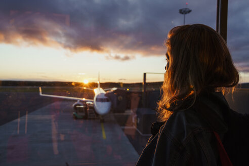 Junge Frau schaut durch das Fenster im Flugzeug auf dem Flughafen bei Sonnenuntergang - KKAF01802
