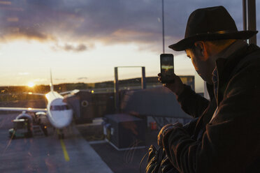 Junger Mann hinter dem Fenster, der mit seinem Handy ein Foto von einem Flugzeug auf dem Flughafen macht - KKAF01803