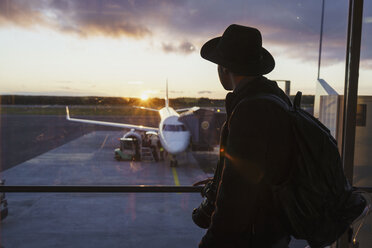 Young man with hat and backpack looking through window on plane at the airport - KKAF01805