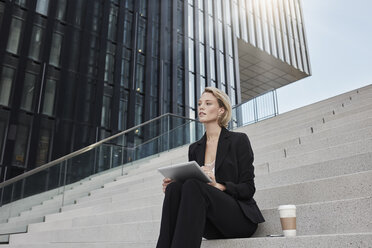 Blonde Geschäftsfrau mit Tablet und Kaffee zum Mitnehmen sitzt auf einer Treppe vor einem modernen Bürogebäude - RORF01478