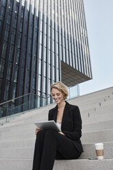 Blonde Geschäftsfrau mit Kaffee zum Mitnehmen sitzt auf einer Treppe vor einem modernen Bürogebäude und benutzt ein Tablet - RORF01479