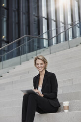 Porträt einer lächelnden Geschäftsfrau mit Tablet und Kaffee zum Mitnehmen, die auf einer Treppe vor einem modernen Bürogebäude sitzt - RORF01480