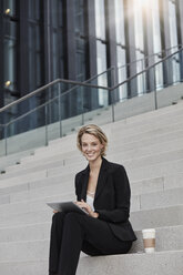 Portrait of smiling businesswoman with tablet and coffee to go sitting on stairs in front of modern office building - RORF01480