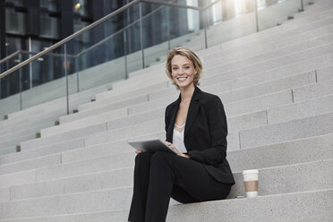 Portrait of smiling businesswoman with tablet and coffee to go sitting on stairs outdoors - RORF01481