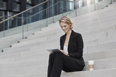 Blonde Geschäftsfrau mit Kaffee zum Mitnehmen, die auf einer Treppe sitzt und ein Tablet benutzt - RORF01482