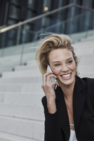 Porträt einer lachenden Geschäftsfrau, die auf einer Treppe im Freien sitzt und telefoniert, lizenzfreies Stockfoto