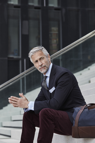 Porträt eines eleganten Geschäftsmannes mit Reisetasche auf einer Treppe sitzend im Freien, lizenzfreies Stockfoto