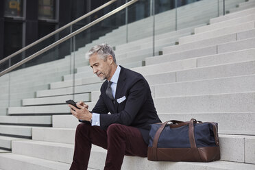 Fashionable businessman with travelling bag sitting on stairs using mobile phone - RORF01492