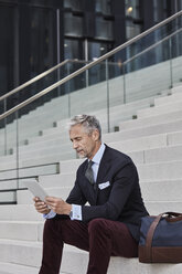 Modischer Geschäftsmann mit Reisetasche, der auf einer Treppe sitzt und ein Tablet benutzt - RORF01494